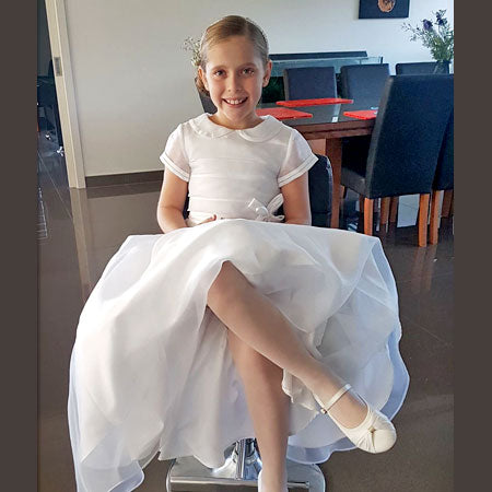 Head to toe photo of smiling girl in white first communion dress, tights and shoes, sitting on chair with her fair hair tied back, dining table and chairs in background.