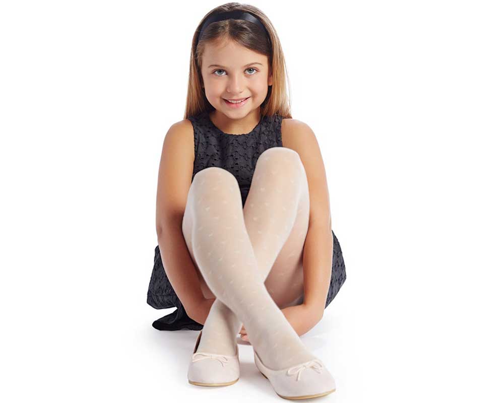 Young girl sitting and smiling as she faces the camera with legs crossed in white sheer tights, white ballet shoes, black dress and black headband in her brown hair.