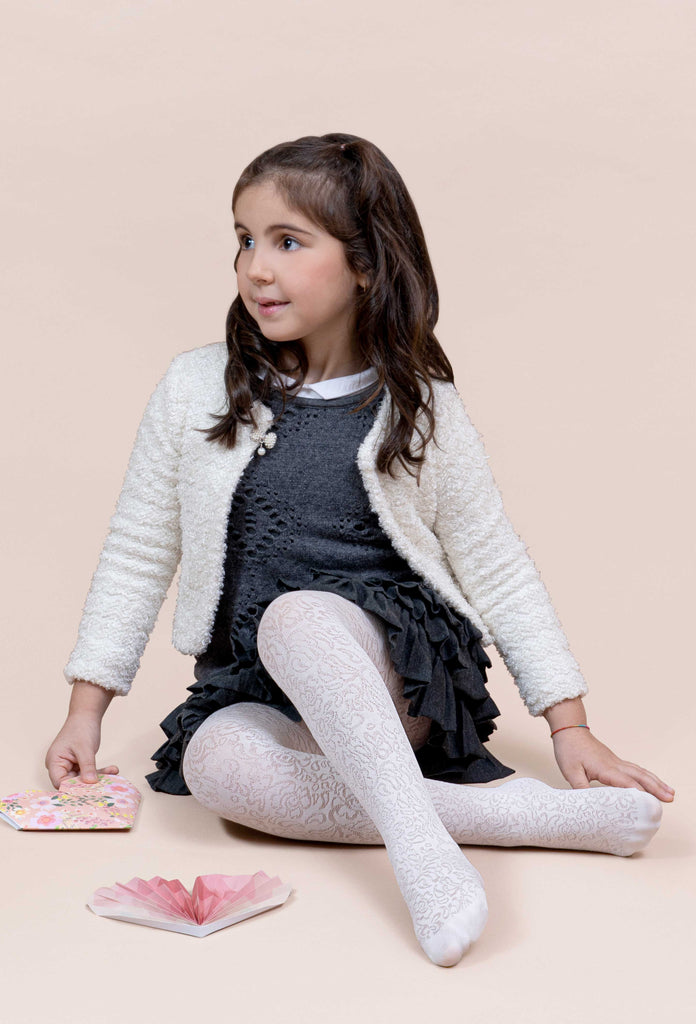 Young girl, sitting on the floor with legs crossed over, in pattern cream tights and black pinafore.