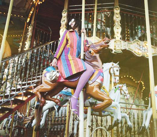 Lady astride a carousel horse wearing brightly coloured clothes.