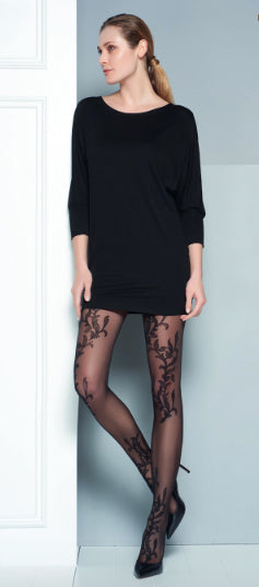 Lady standing against white door wearing a long black top and Mariel tights which are patterned with a vine leaf that is repeated throughout this black sheer tight