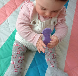 Top down view of baby from forehead to toes, baby wearing pink ruffled top, white knit overalls and grey tights with hearts, sitting on striped colored blanket and holding purple teething toy.