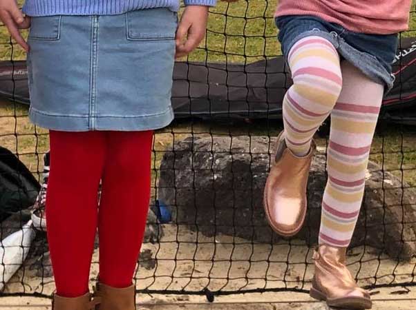 Two young girls on climbing net at park, close-up shot of legs. One girl in blue denim skirt and red winter tights, one girl in denim skirt, multi colored striped tights and shiny ankle boots.