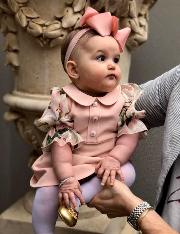 Adorable baby girl of Middle Eastern heritage dressed in special occasion pink dress with floral puff sleeves, lavender tights, gold shoes and a big pink bow around her head, large classical garden urn in background.