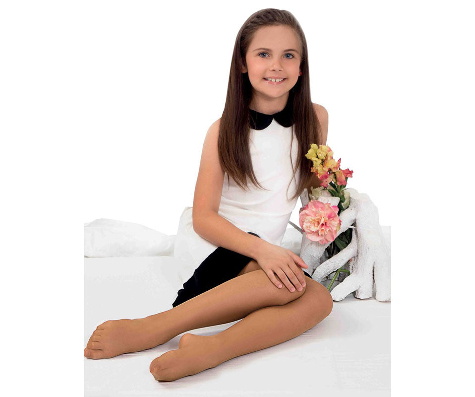 Head to toes of young girl smiling as she sits in black and white special occasion dress and skin toned tights, next to a vase of flowers.
