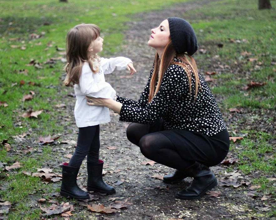 Close up of mother crouching down to talk to her small daughter in park on wet day. Woman is in black with black hat, girl is in black boots and leggings with white top. Autumn leaves scattered in background.