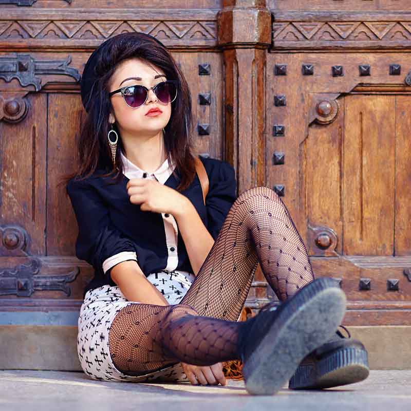 Stylish dark haired woman in black and white outfit, black net tights, sunglasses and lace up shoes sitting against old church doors in a carefree fashion.