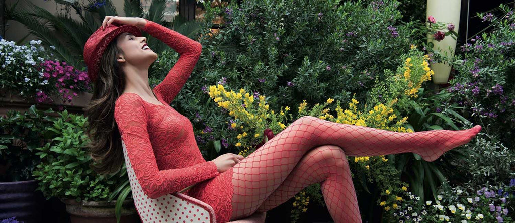 Profile view of tall young woman in short red lace dress, red fishnet tights and red straw hat, laughing and reclining in outdoor chair in courtyard filled with abundant, multi-colored flowering plants.
