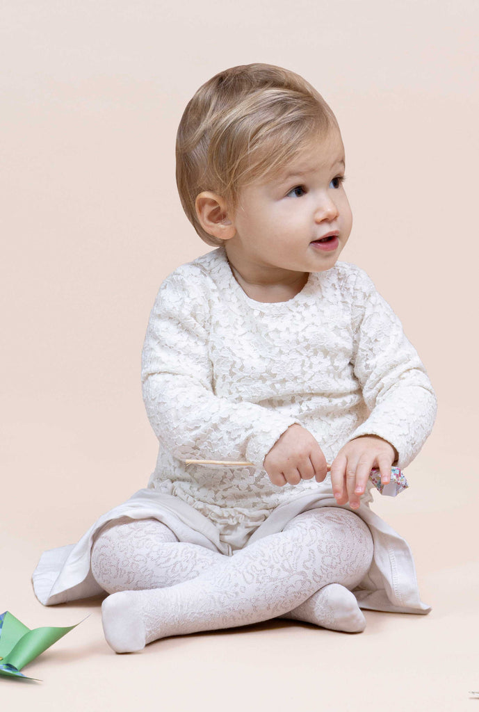 Baby sitting with legs crossed, wearing white dress and lace tights.