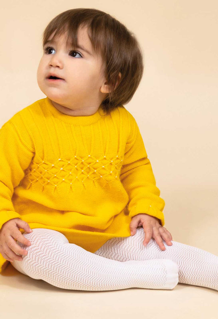 Baby girl, sitting , wearing a yellow knit dress and white patterned tights.