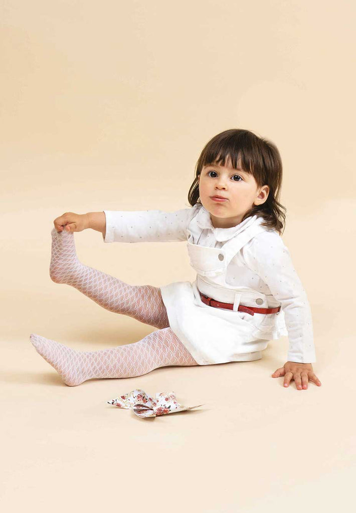 Toddler holding on to her foot, wearing white tights and pinafore.